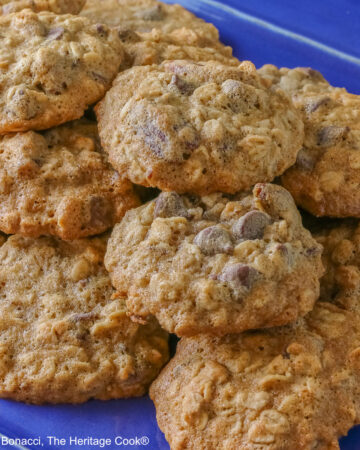 A large pile of amazing, delicious Chewy Oatmeal Chocolate Chip Cookies stacked on a rectangular blue platter sitting on a mottled gray background © 2026 Jane Bonacci, The Heritage Cook.