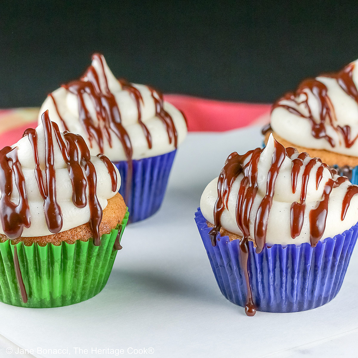 Dalmatian Cupcakes in a variety of boldly colored liners topped with mounds of white frosting and drizzled with chocolate ganache, sitting on a white surface with a black background © 2025 Jane Bonacci, The Heritage Cook. 