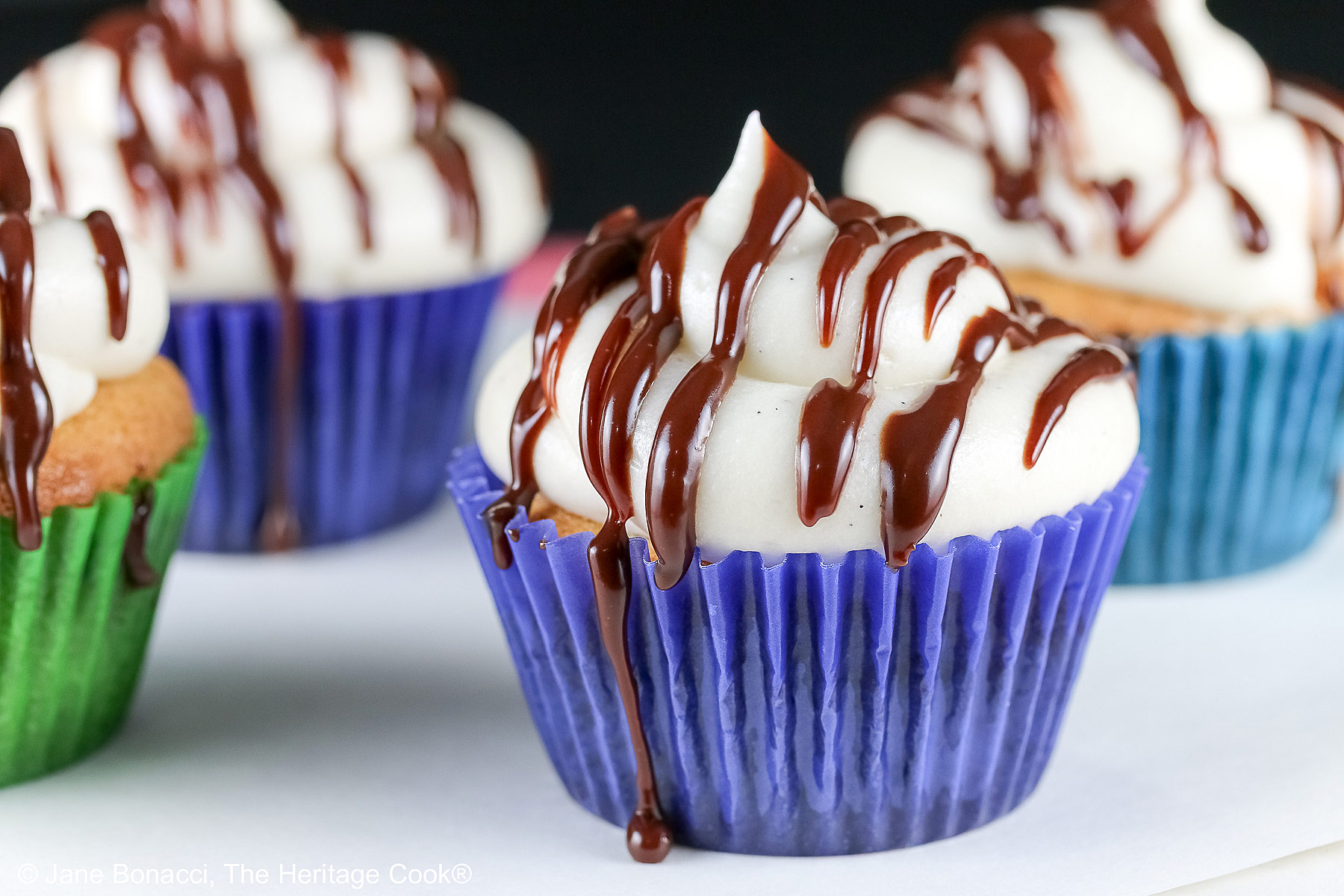Dalmatian Cupcakes in a variety of boldly colored liners topped with mounds of white frosting and drizzled with chocolate ganache, sitting on a white surface with a black background © 2025 Jane Bonacci, The Heritage Cook. 