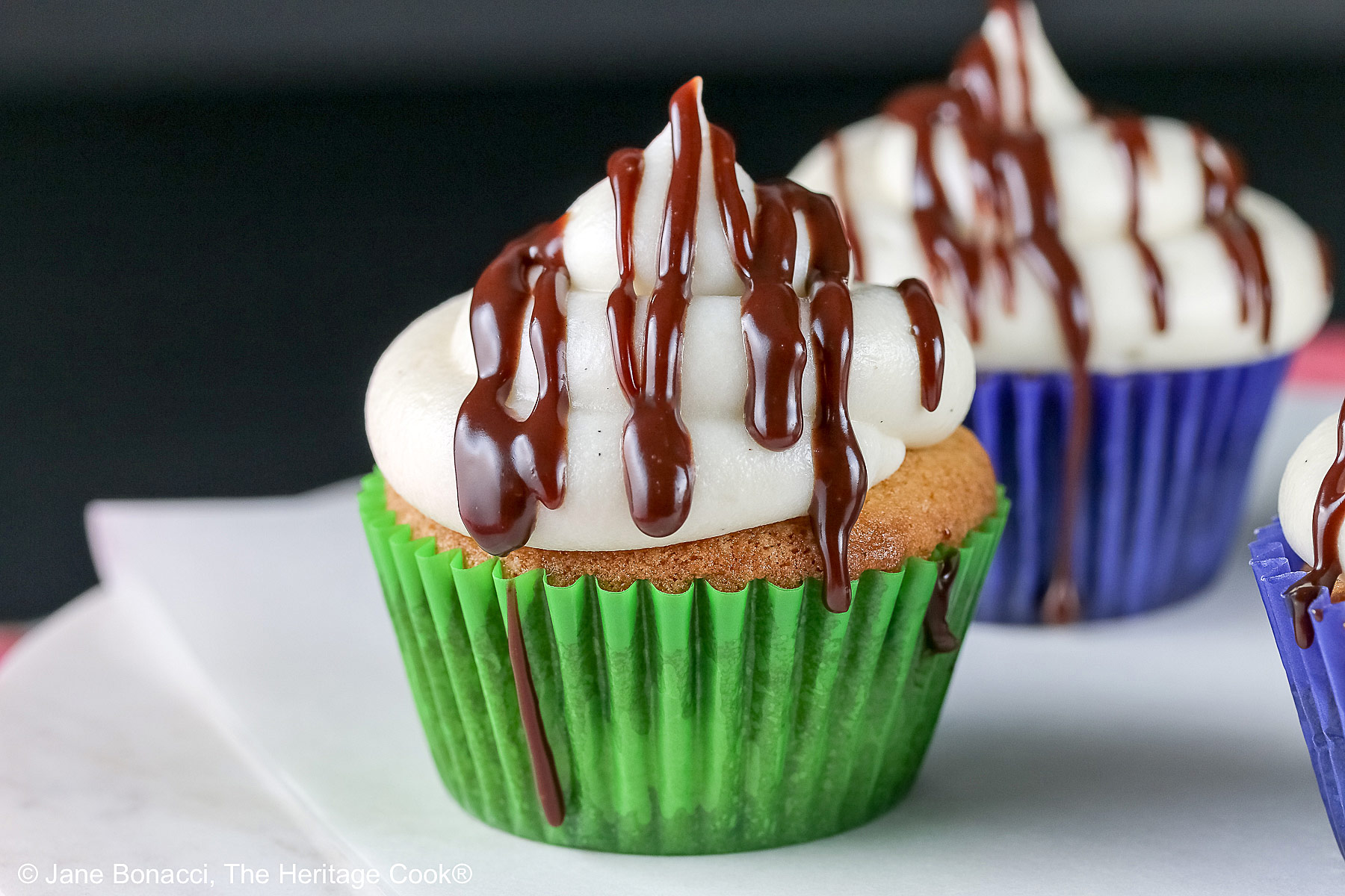 Dalmatian Cupcakes in a variety of boldly colored liners topped with mounds of white frosting and drizzled with chocolate ganache, sitting on a white surface with a black background © 2025 Jane Bonacci, The Heritage Cook. 