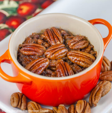 Line of ramekins and cocottes in a variety of colors on a white tray filled with crustless bourbon pecan pies with pecans scattered around, sitting on a cloth covered with red apples and green leaves © 2025 Jane Bonacci, The Heritage Cook.