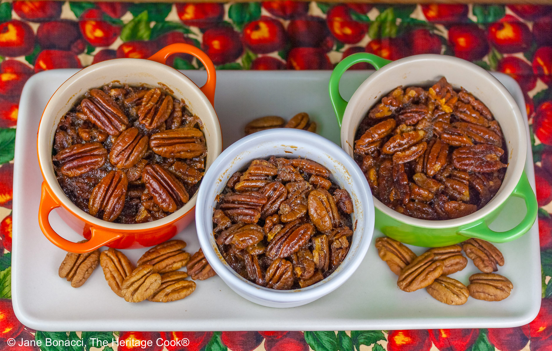 Line of ramekins and cocottes in a variety of colors on a white tray filled with crustless bourbon pecan pies with pecans scattered around, sitting on a cloth covered with red apples and green leaves © 2025 Jane Bonacci, The Heritage Cook.