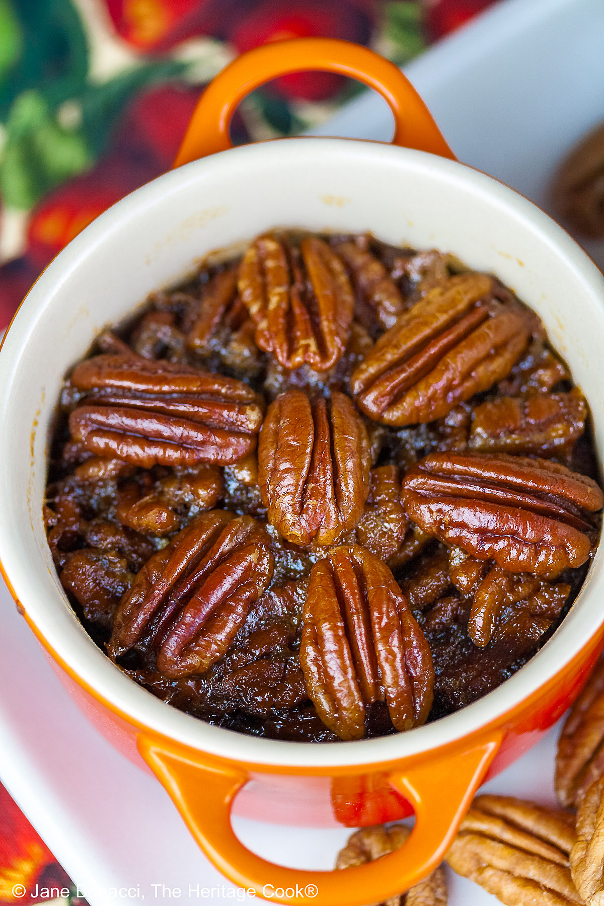 Line of ramekins and cocottes in a variety of colors on a white tray filled with crustless bourbon pecan pies with pecans scattered around, sitting on a cloth covered with red apples and green leaves © 2025 Jane Bonacci, The Heritage Cook.