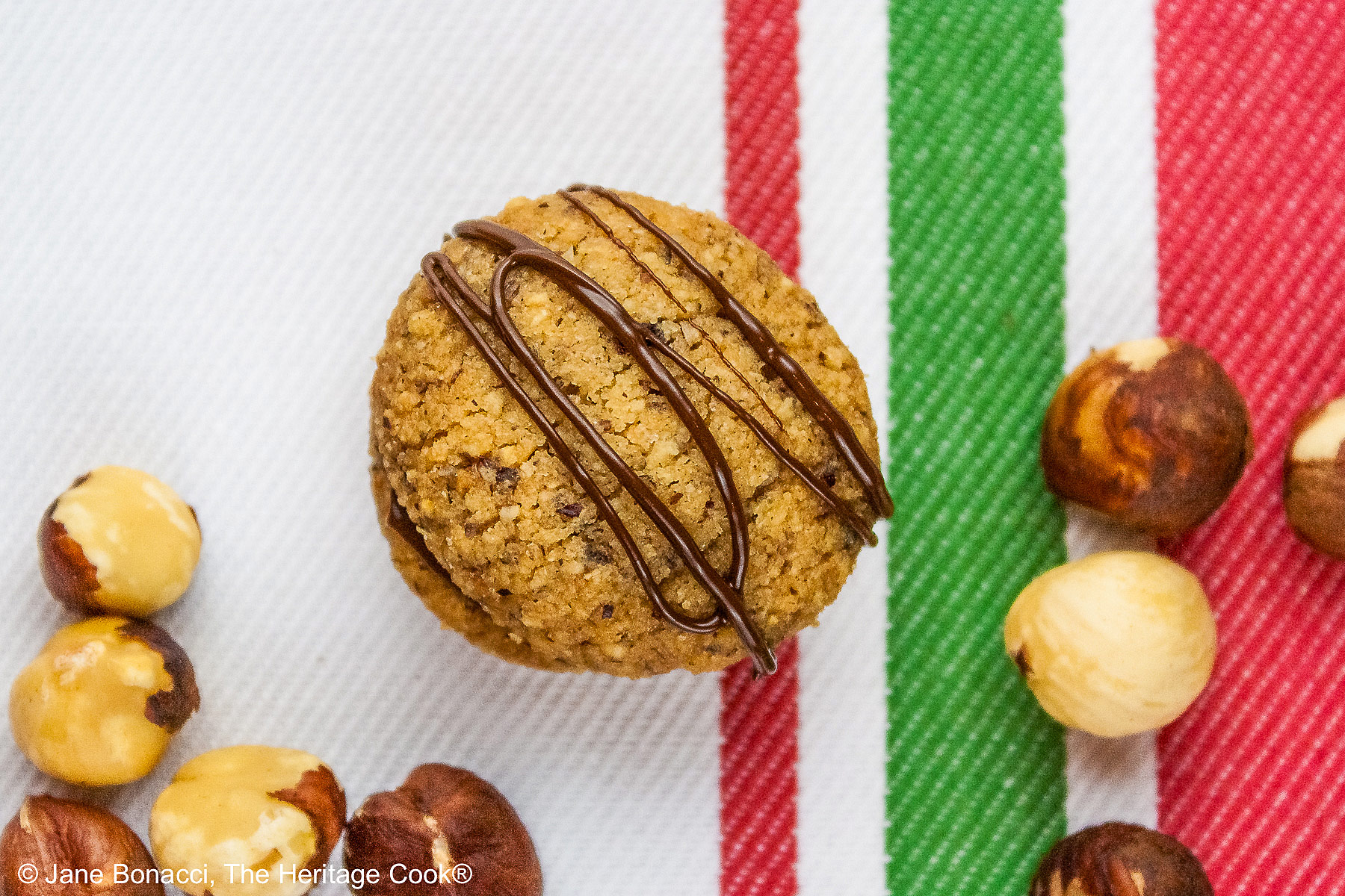 Chocolate Hazelnut Sandwich Cookies filled with chocolate ganache on a white plate sitting on a red, white, and green Italian towel, some drizzled with melted chocolate, with hazelnuts scattered around the cookies © 2026 Jane Bonacci, The Heritage Cook.