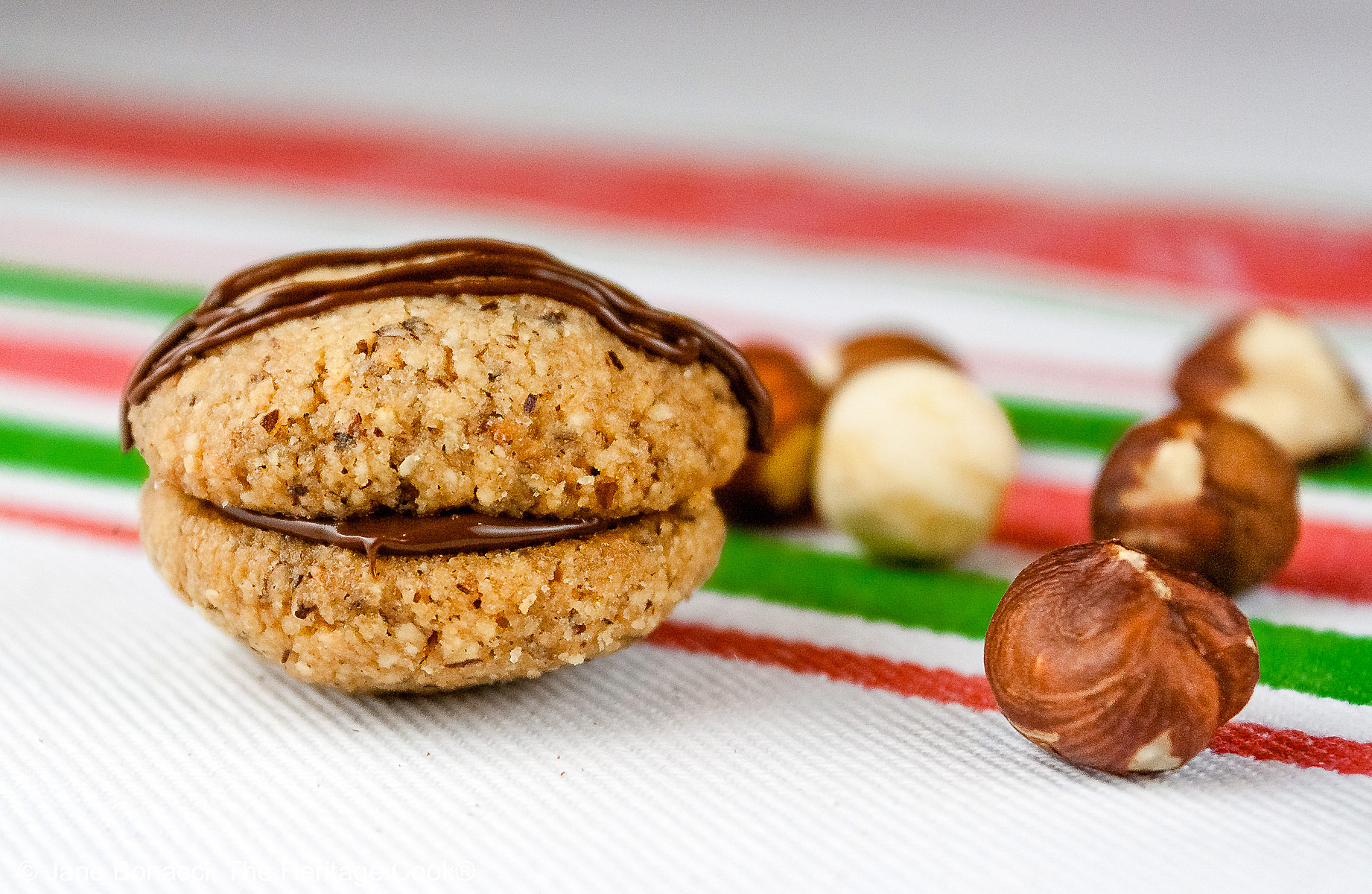 Chocolate Hazelnut Sandwich Cookies filled with chocolate ganache on a white plate sitting on a red, white, and green Italian towel, some drizzled with melted chocolate, with hazelnuts scattered around the cookies © 2026 Jane Bonacci, The Heritage Cook.