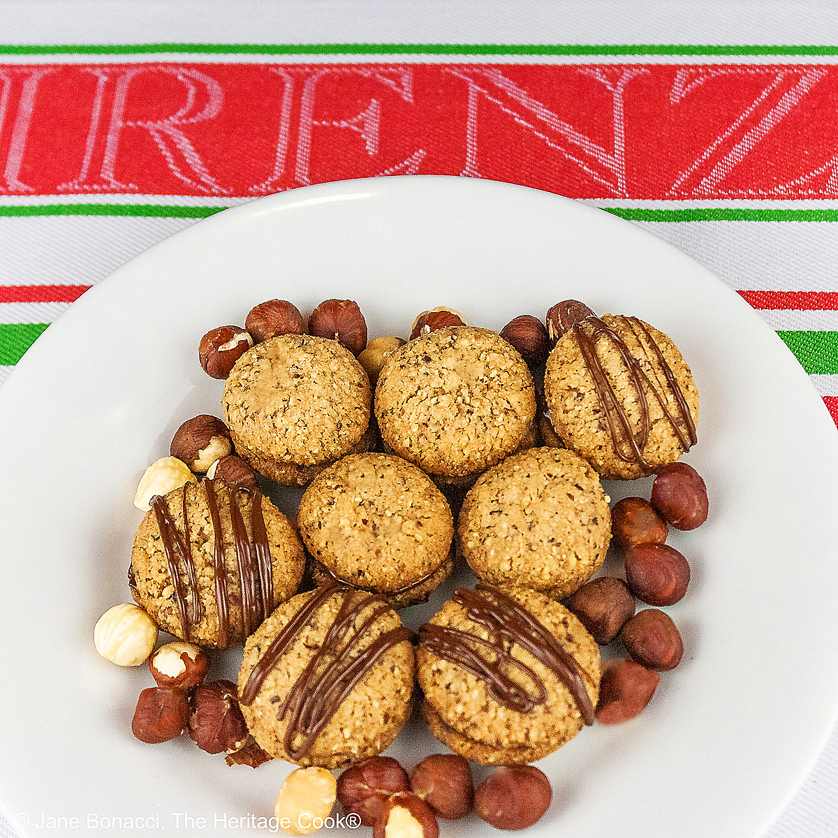 Chocolate Hazelnut Sandwich Cookies filled with chocolate ganache on a white plate sitting on a red, white, and green Italian towel, some drizzled with melted chocolate, with hazelnuts scattered around the cookies © 2026 Jane Bonacci, The Heritage Cook.