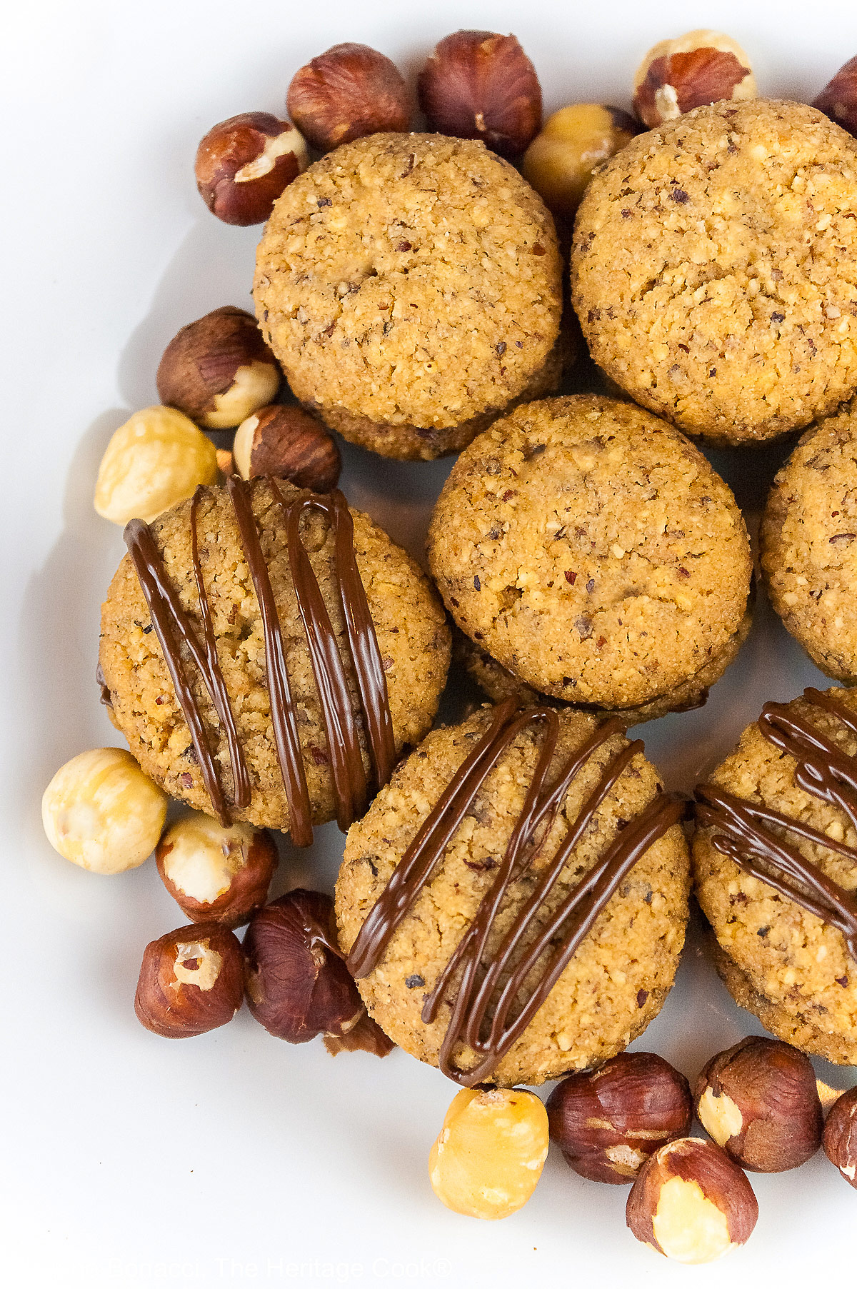 Chocolate Hazelnut Sandwich Cookies filled with chocolate ganache on a white plate sitting on a red, white, and green Italian towel, some drizzled with melted chocolate, with hazelnuts scattered around the cookies © 2026 Jane Bonacci, The Heritage Cook.