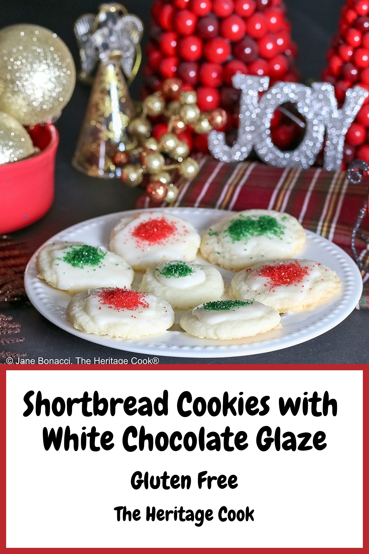 Plate full of shortbread cookies topped with a white chocolate glaze and sprinkled with red and green sugars, sitting on a white plate with holiday decorations like gold ornaments, red trees, a mini red sleigh, and seasonal placemats © 2025 Jane Bonacci, The Heritage Cook.