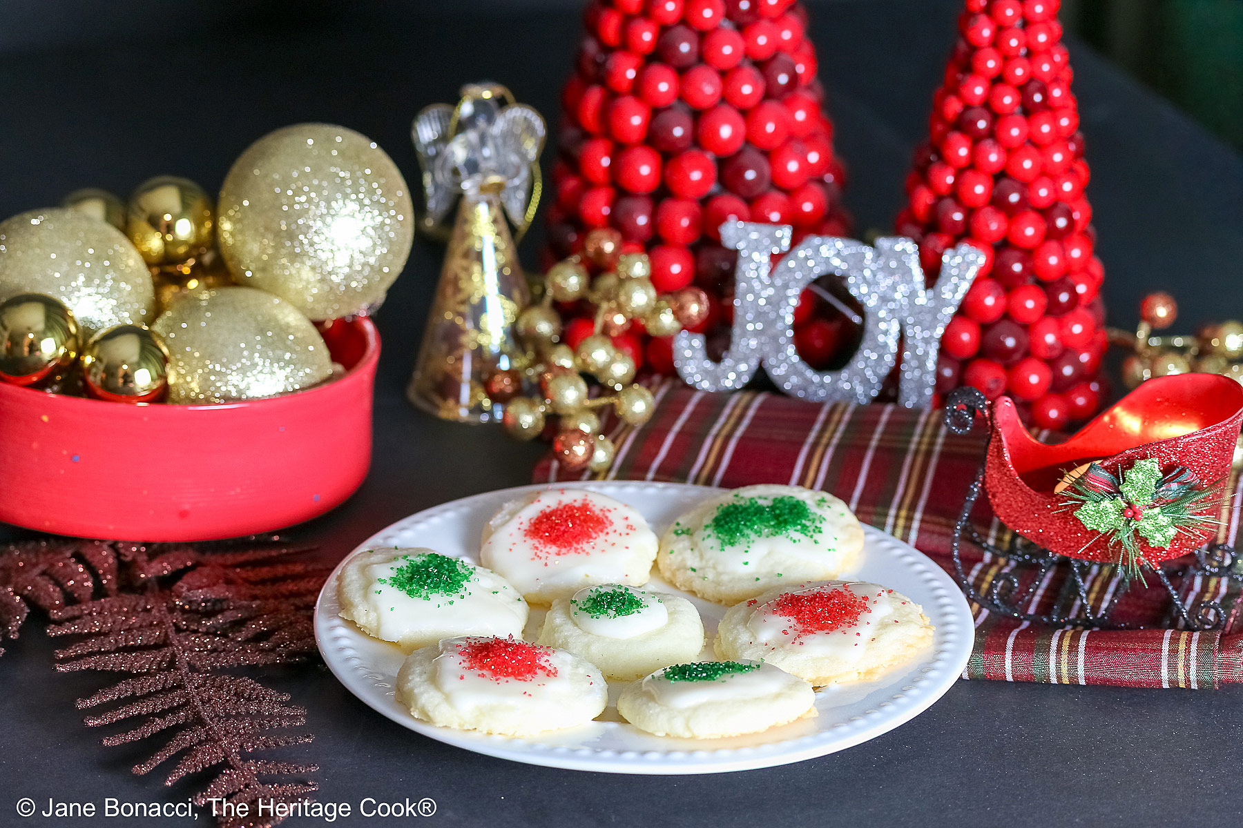 Plate full of shortbread cookies topped with a white chocolate glaze and sprinkled with red and green sugars, sitting on a white plate with holiday decorations like gold ornaments, red trees, a mini red sleigh, and seasonal placemats © 2025 Jane Bonacci, The Heritage Cook.