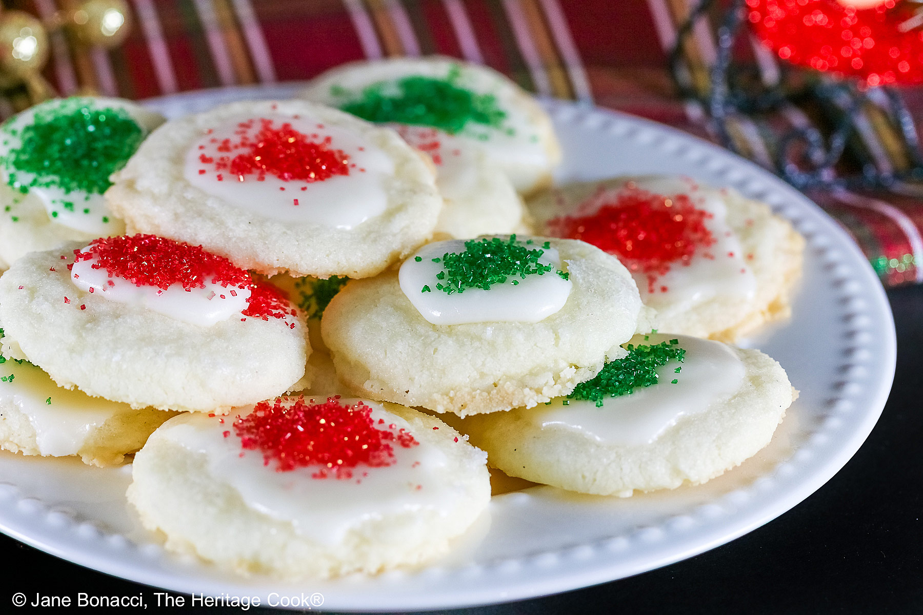 Plate full of shortbread cookies topped with a white chocolate glaze and sprinkled with red and green sugars, sitting on a white plate with holiday decorations like gold ornaments, red trees, a mini red sleigh, and seasonal placemats © 2025 Jane Bonacci, The Heritage Cook.