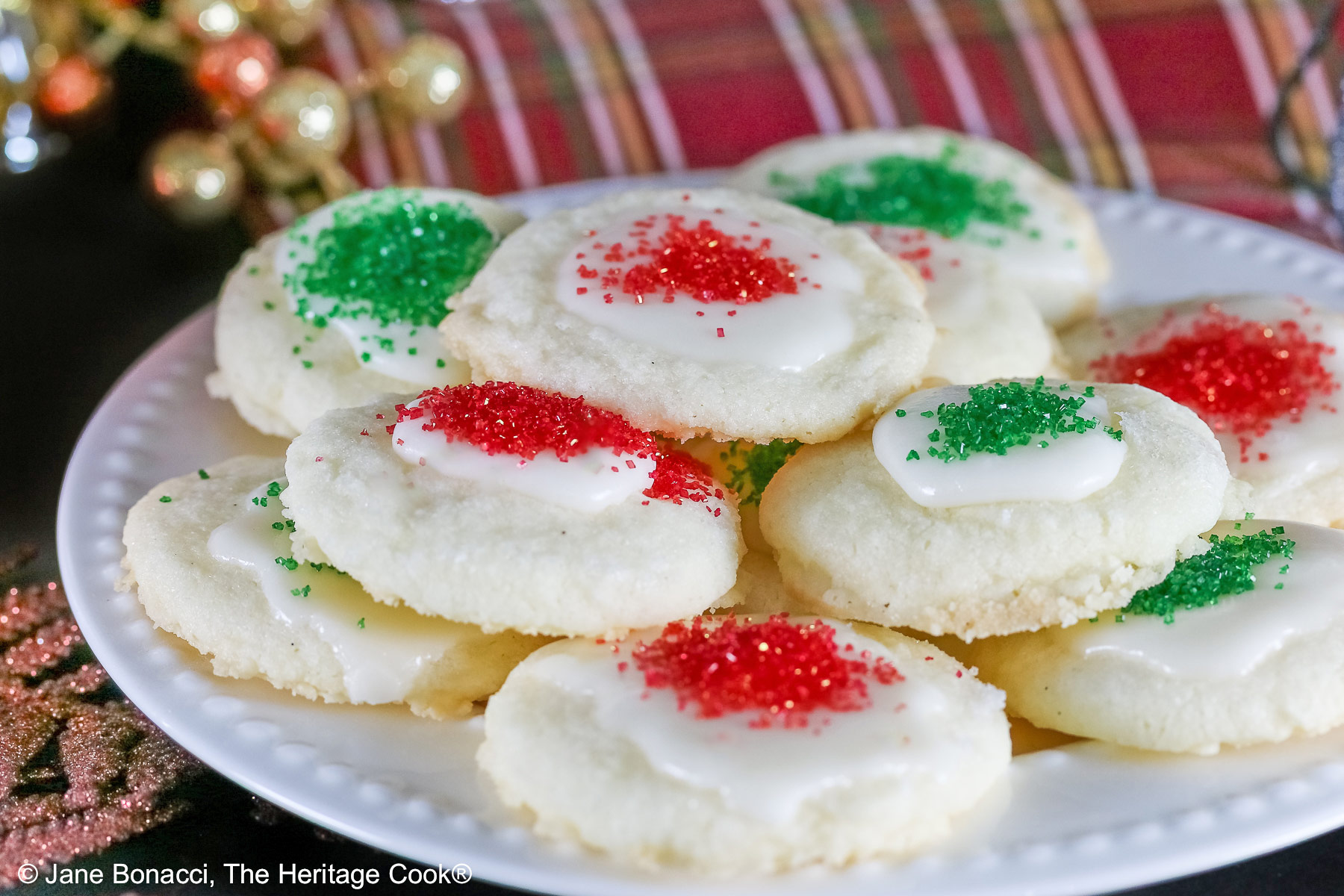 Plate full of shortbread cookies topped with a white chocolate glaze and sprinkled with red and green sugars, sitting on a white plate with holiday decorations like gold ornaments, red trees, a mini red sleigh, and seasonal placemats © 2025 Jane Bonacci, The Heritage Cook.