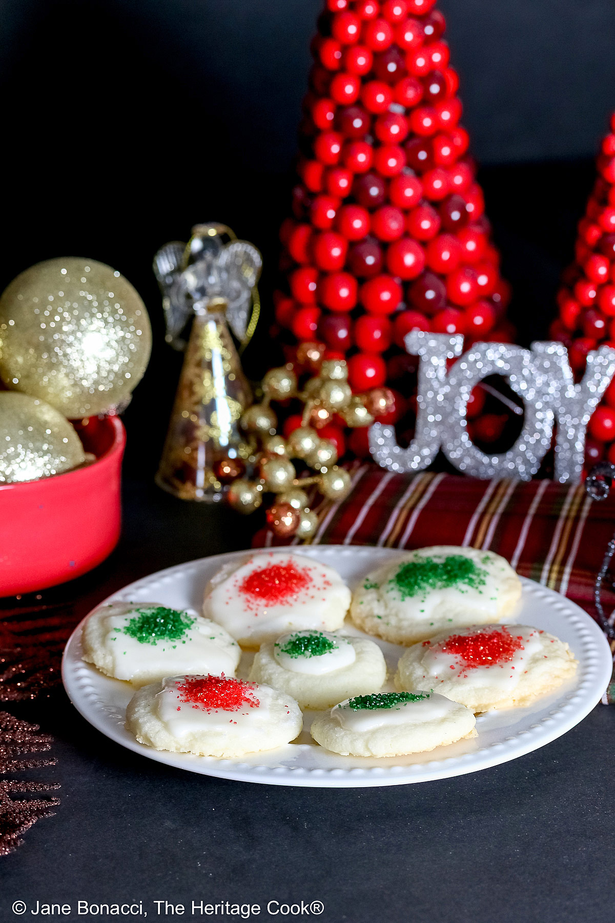 Plate full of shortbread cookies topped with a white chocolate glaze and sprinkled with red and green sugars, sitting on a white plate with holiday decorations like gold ornaments, red trees, a mini red sleigh, and seasonal placemats © 2025 Jane Bonacci, The Heritage Cook.