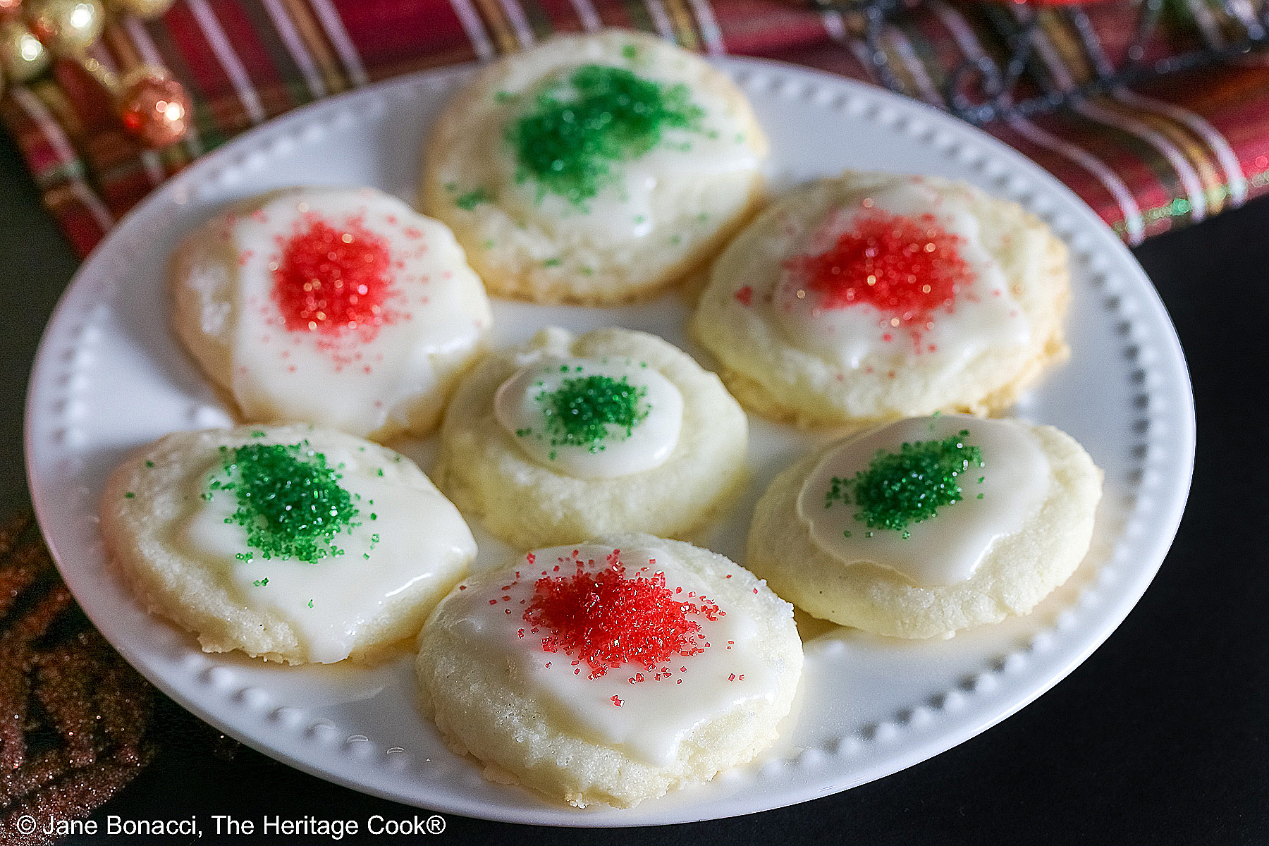 Plate full of shortbread cookies topped with a white chocolate glaze and sprinkled with red and green sugars, sitting on a white plate with holiday decorations like gold ornaments, red trees, a mini red sleigh, and seasonal placemats © 2025 Jane Bonacci, The Heritage Cook.