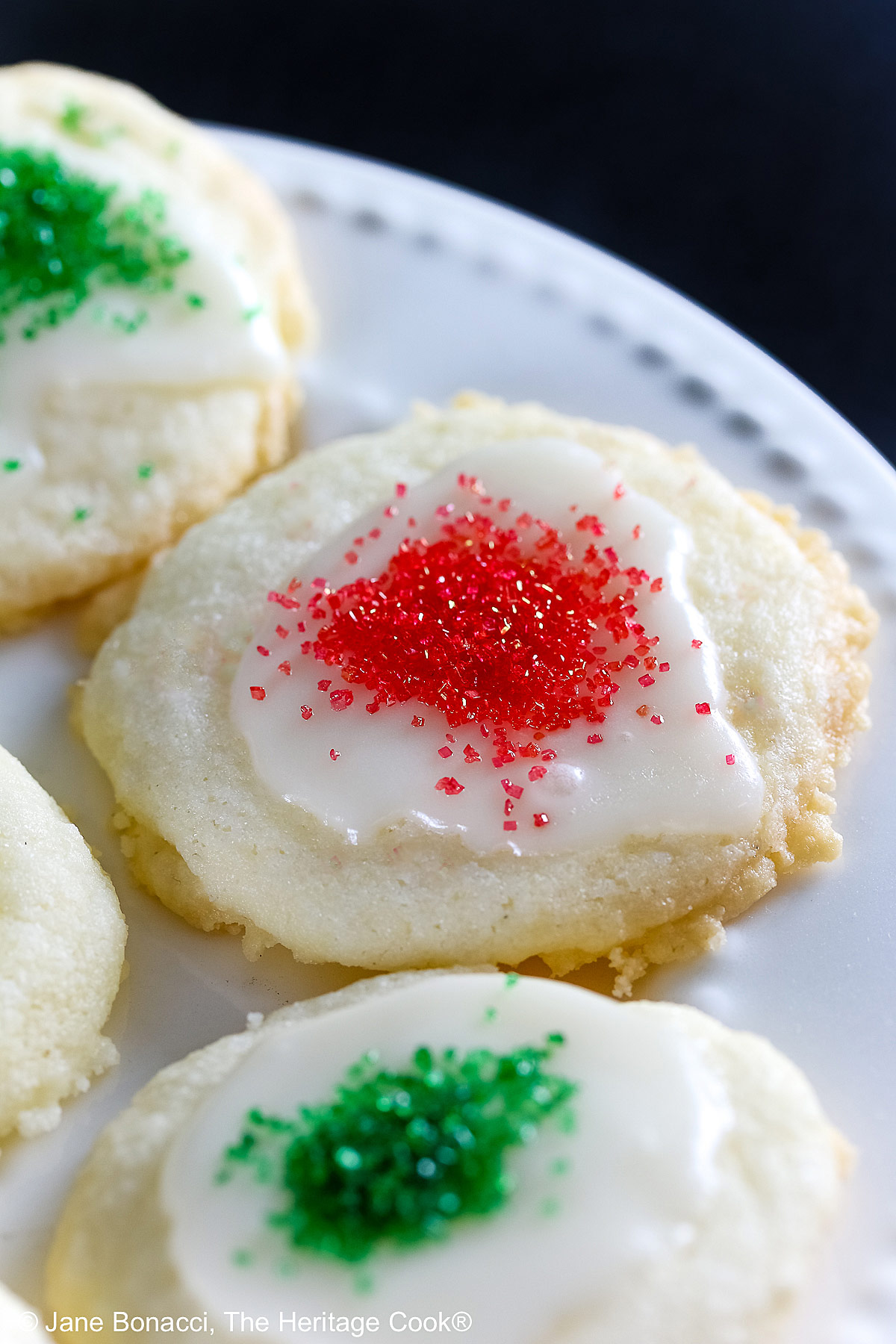 Plate full of shortbread cookies topped with a white chocolate glaze and sprinkled with red and green sugars, sitting on a white plate with holiday decorations like gold ornaments, red trees, a mini red sleigh, and seasonal placemats © 2025 Jane Bonacci, The Heritage Cook.