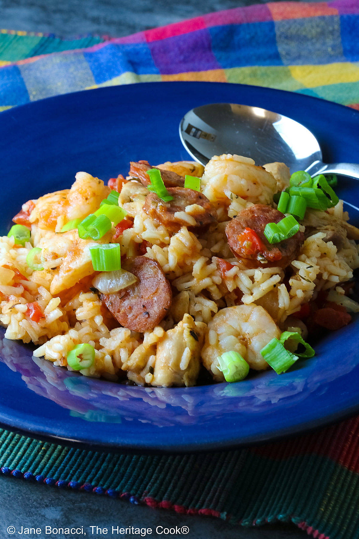 Mound of Easy New Orleans Jambalaya with chicken, shrimp, and sausages prominently placed and sprinkled with sliced green onions all in a dark blue bowl sitting on a brightly colored checked cloth © 2026 Jane Bonacci, The Heritage Cook.