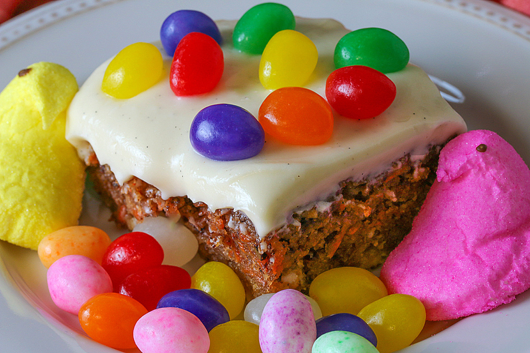 Square slice of White Chocolate Carrot Cake on a cream colored plate, surrounded by and topped with brightly colored jelly beans and marshmallow Peeps on a deep pink cloth © 2024 Jane Bonacci, The Heritage Cook.