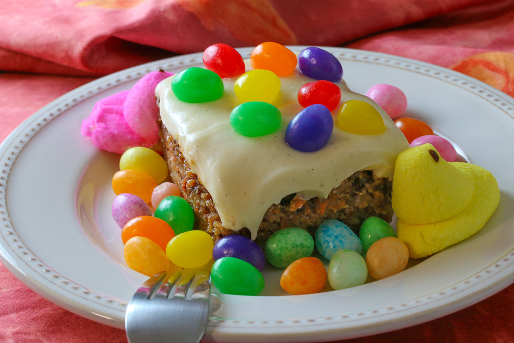 Square slice of White Chocolate Carrot Cake on a cream colored plate, surrounded by and topped with brightly colored jelly beans and marshmallow Peeps on a deep pink cloth © 2024 Jane Bonacci, The Heritage Cook.