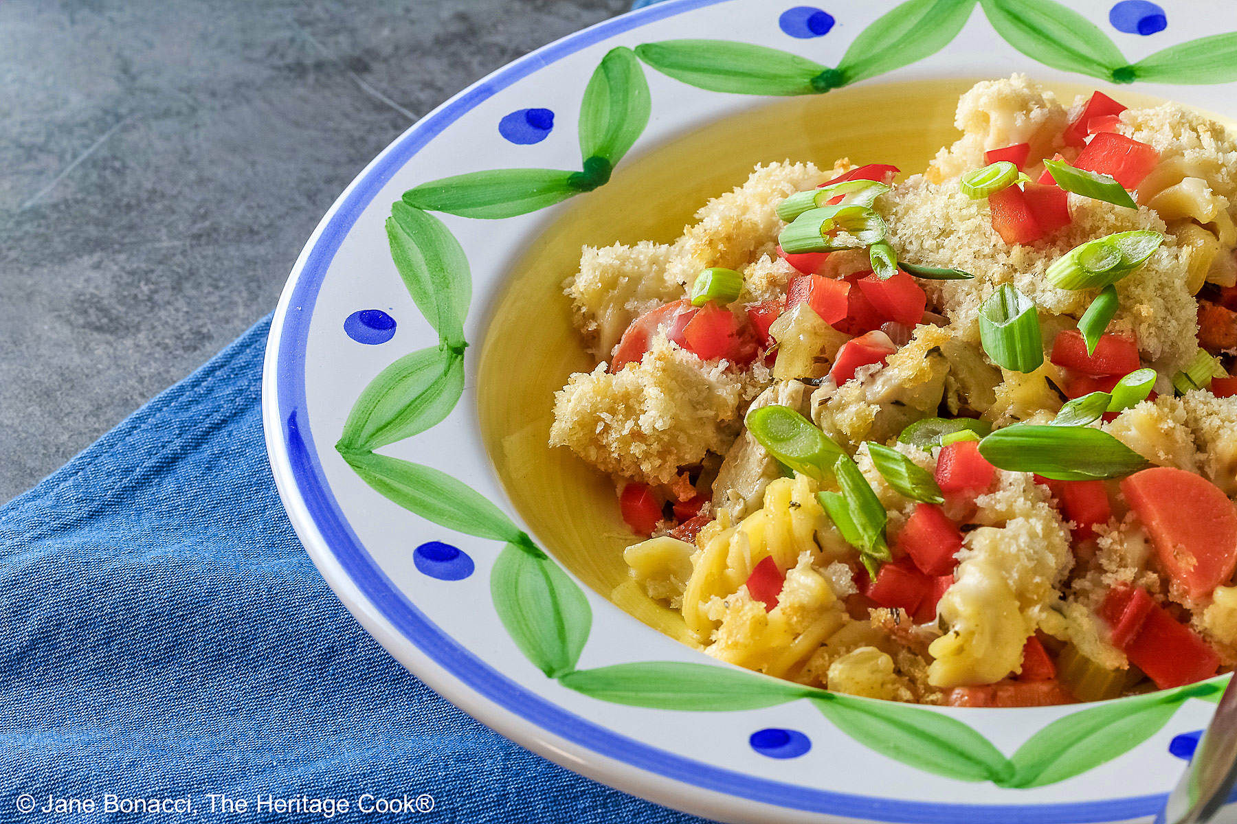 Bowl with a large rim decorated with green and blue designs filled with the Chicken Pot Pie Pasta Casserole that is topped with tiny squares of red bell pepper and sliced green onions on a blue denim cloth © 2026 Jane Bonacci, The Heritage Cook.
