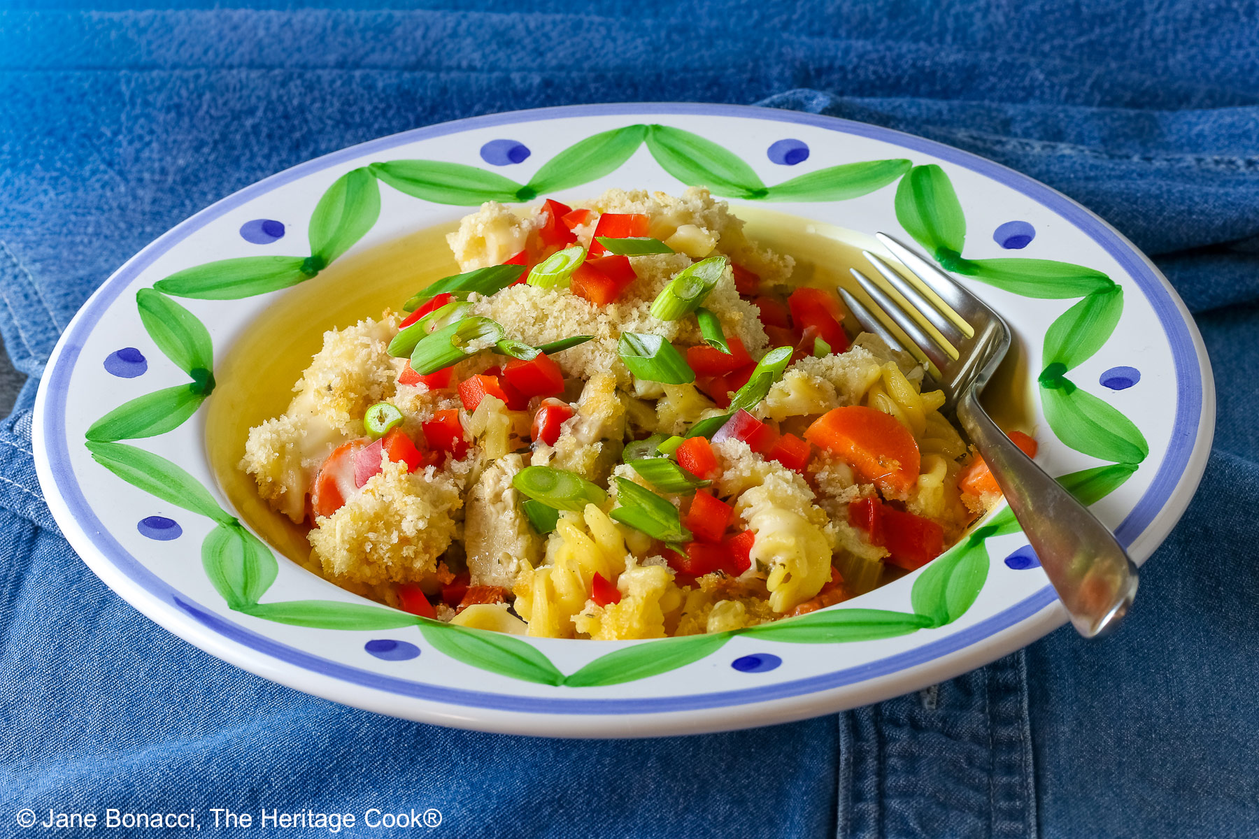 Bowl with a large rim decorated with green and blue designs filled with the Chicken Pot Pie Pasta Casserole that is topped with tiny squares of red bell pepper and sliced green onions on a blue denim cloth © 2026 Jane Bonacci, The Heritage Cook.