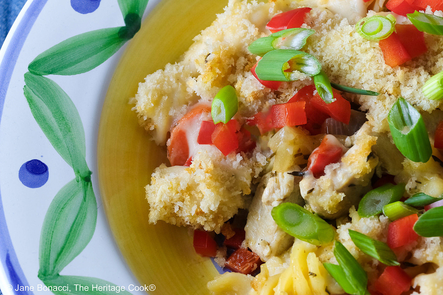 Bowl with a large rim decorated with green and blue designs filled with the Chicken Pot Pie Pasta Casserole that is topped with tiny squares of red bell pepper and sliced green onions on a blue denim cloth © 2026 Jane Bonacci, The Heritage Cook.