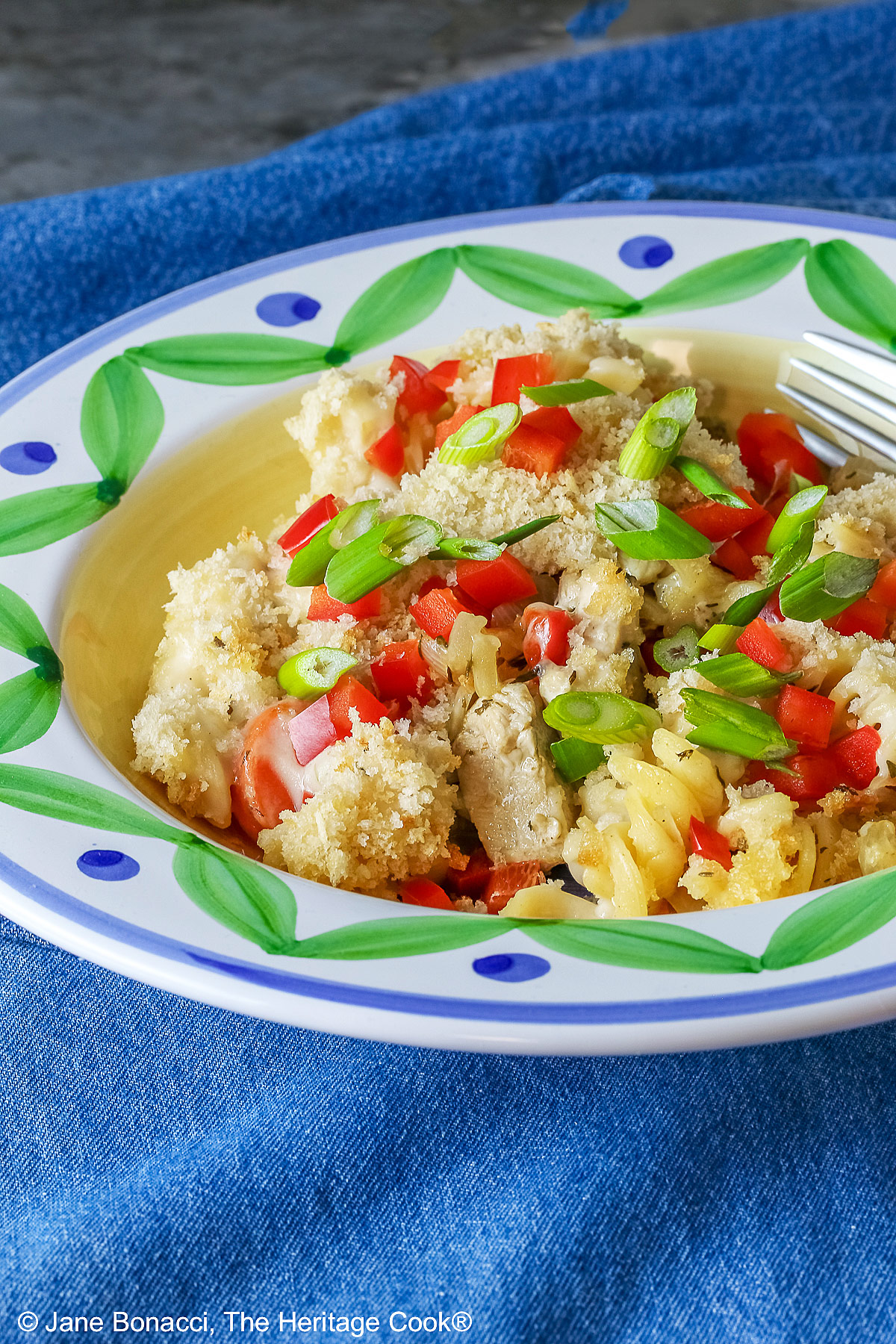 Bowl with a large rim decorated with green and blue designs filled with the Chicken Pot Pie Pasta Casserole that is topped with tiny squares of red bell pepper and sliced green onions on a blue denim cloth © 2026 Jane Bonacci, The Heritage Cook.