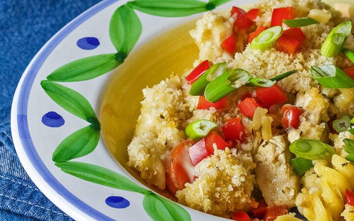 Bowl with a large rim decorated with green and blue designs filled with the Chicken Pot Pie Pasta Casserole that is topped with tiny squares of red bell pepper and sliced green onions on a blue denim cloth © 2026 Jane Bonacci, The Heritage Cook.