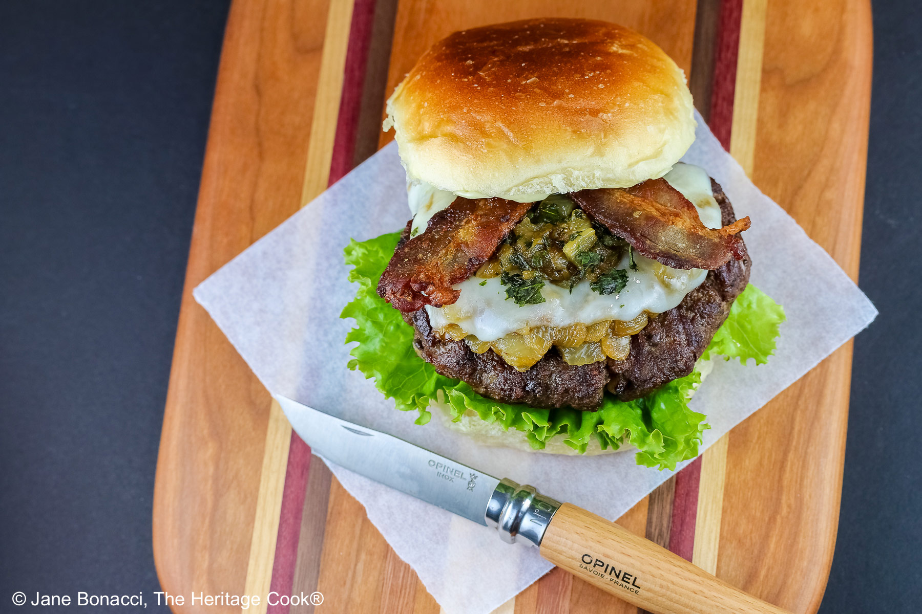 Grilled Santa Fe Green Chile Bacon Burger sitting on a square of parchment on top of a beautiful striped cutting board with lettuce on the bottom bun beneath the burger patty, with sauteed onions and green chiles, melted cheese, and strips of bacon © 2026 Jane Bonacci, The Heritage Cook.
