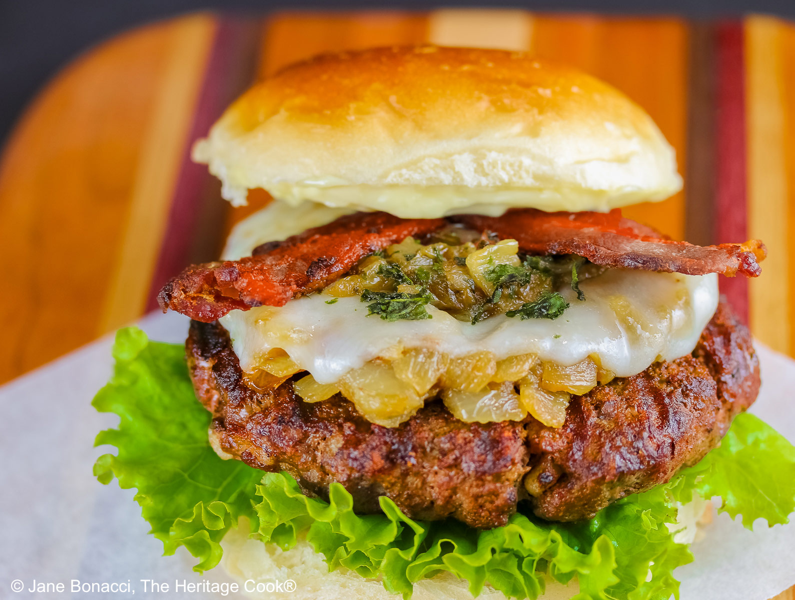 Grilled Santa Fe Green Chile Bacon Burger sitting on a square of parchment on top of a beautiful striped cutting board with lettuce on the bottom bun beneath the burger patty, with sauteed onions and green chiles, melted cheese, and strips of bacon © 2026 Jane Bonacci, The Heritage Cook.