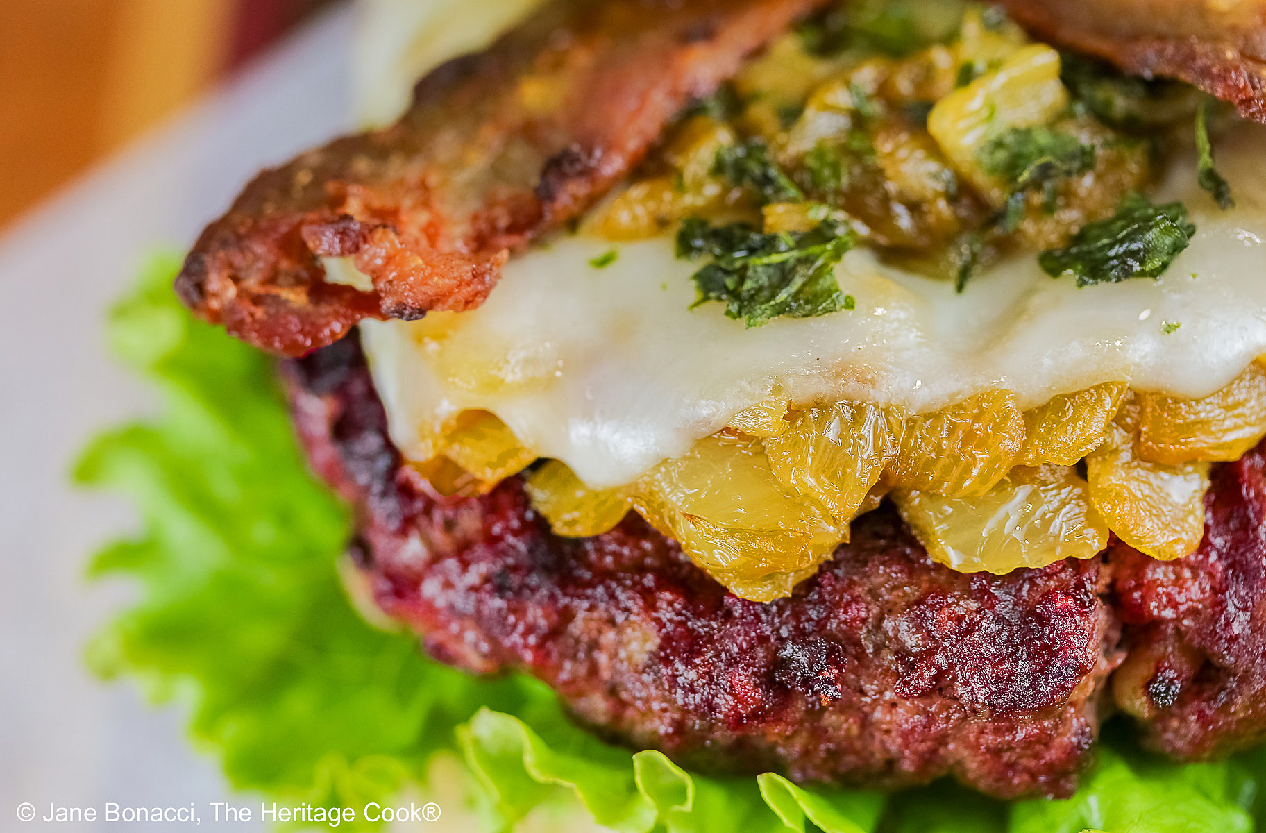 Grilled Santa Fe Green Chile Bacon Burger sitting on a square of parchment on top of a beautiful striped cutting board with lettuce on the bottom bun beneath the burger patty, with sauteed onions and green chiles, melted cheese, and strips of bacon © 2026 Jane Bonacci, The Heritage Cook.