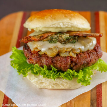 Grilled Santa Fe Green Chile Bacon Burger sitting on a square of parchment on top of a beautiful striped cutting board with lettuce on the bottom bun beneath the burger patty, with sauteed onions and green chiles, melted cheese, and strips of bacon © 2026 Jane Bonacci, The Heritage Cook.