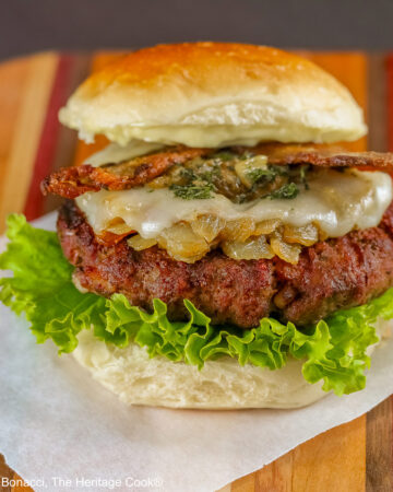 Grilled Santa Fe Green Chile Bacon Burger sitting on a square of parchment on top of a beautiful striped cutting board with lettuce on the bottom bun beneath the burger patty, with sauteed onions and green chiles, melted cheese, and strips of bacon © 2026 Jane Bonacci, The Heritage Cook.