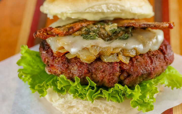 Grilled Santa Fe Green Chile Bacon Burger sitting on a square of parchment on top of a beautiful striped cutting board with lettuce on the bottom bun beneath the burger patty, with sauteed onions and green chiles, melted cheese, and strips of bacon © 2026 Jane Bonacci, The Heritage Cook.