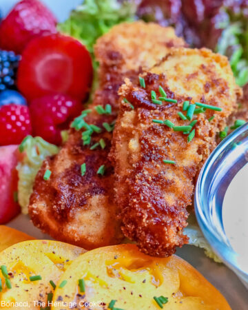 Pile of Buttermilk Fried Chicken Fingers sitting on a green lettuce leaf, on a turquoise plate surrounded by red and blue berries and slices of yellow tomato with a bowl of ranch dressing for dipping, on a black background © 2025 Jane Bonacci, The Heritage Cook.