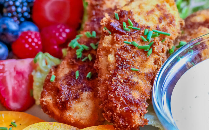 Pile of Buttermilk Fried Chicken Fingers sitting on a green lettuce leaf, on a turquoise plate surrounded by red and blue berries and slices of yellow tomato with a bowl of ranch dressing for dipping, on a black background © 2025 Jane Bonacci, The Heritage Cook.