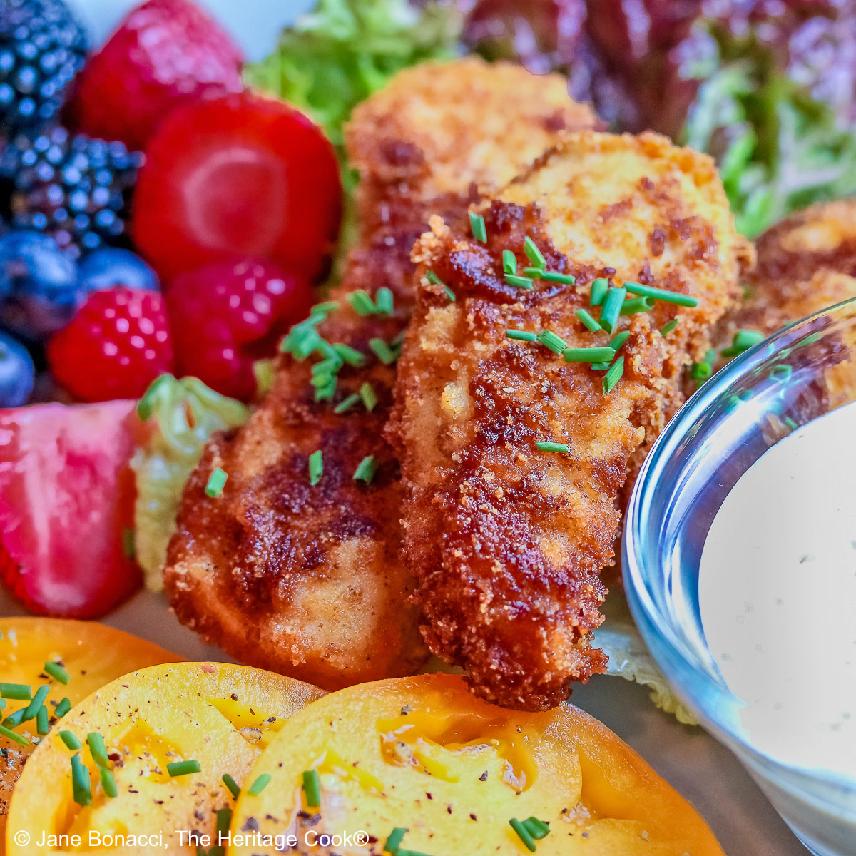 Pile of Buttermilk Fried Chicken Fingers sitting on a green lettuce leaf, on a turquoise plate surrounded by red and blue berries and slices of yellow tomato with a bowl of ranch dressing for dipping, on a black background © 2025 Jane Bonacci, The Heritage Cook.