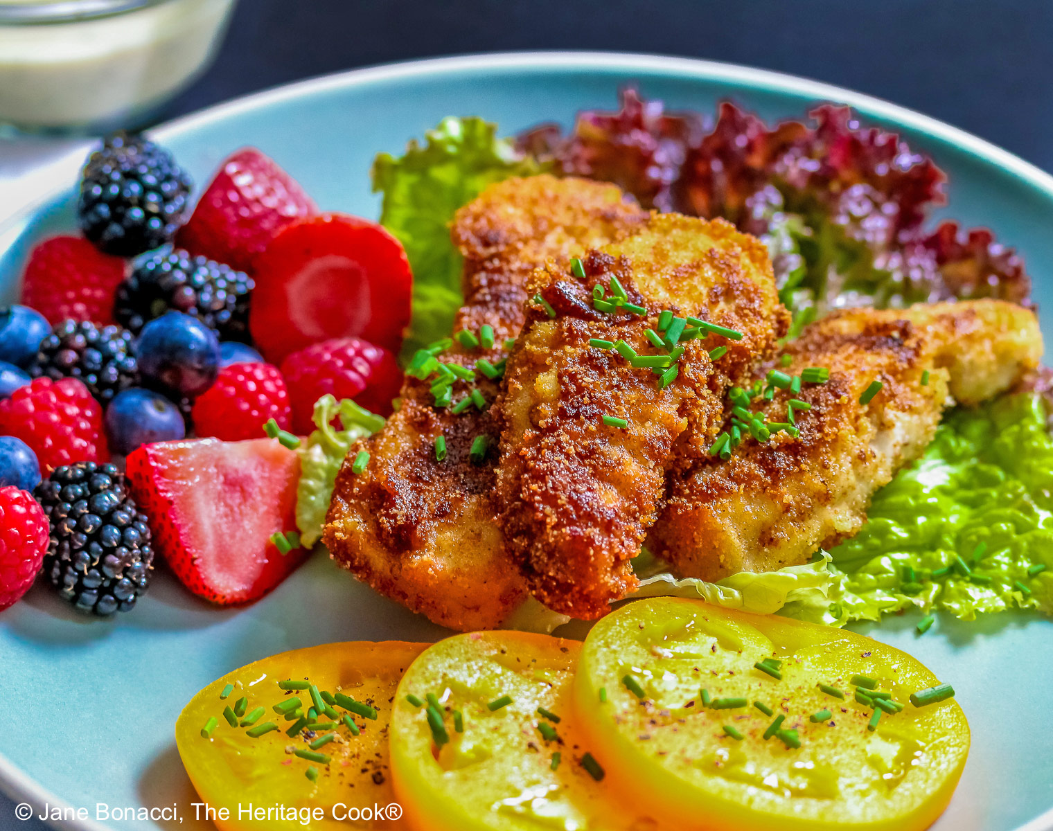 Pile of Buttermilk Fried Chicken Fingers sitting on a green lettuce leaf, on a turquoise plate surrounded by red and blue berries and slices of yellow tomato with a bowl of ranch dressing for dipping, on a black background © 2025 Jane Bonacci, The Heritage Cook. 