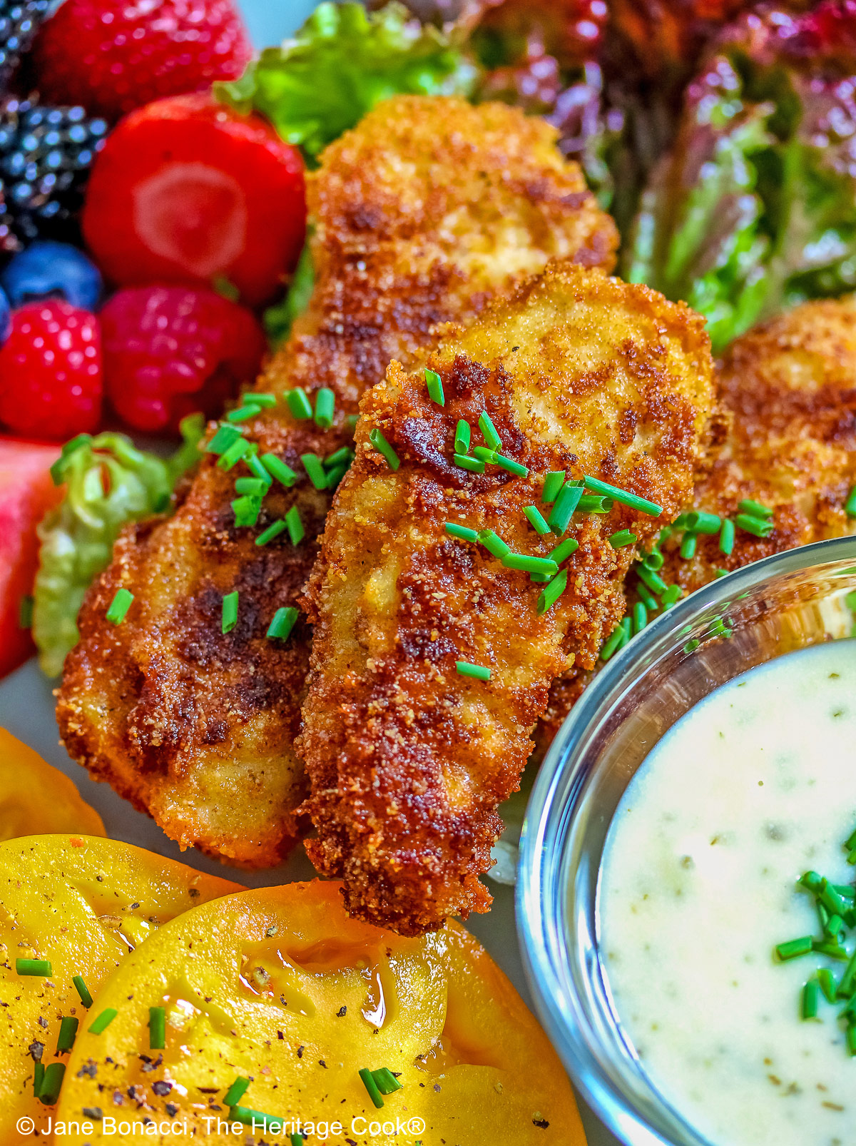 Pile of Buttermilk Fried Chicken Fingers sitting on a green lettuce leaf, on a turquoise plate surrounded by red and blue berries and slices of yellow tomato with a bowl of ranch dressing for dipping, on a black background © 2025 Jane Bonacci, The Heritage Cook. 