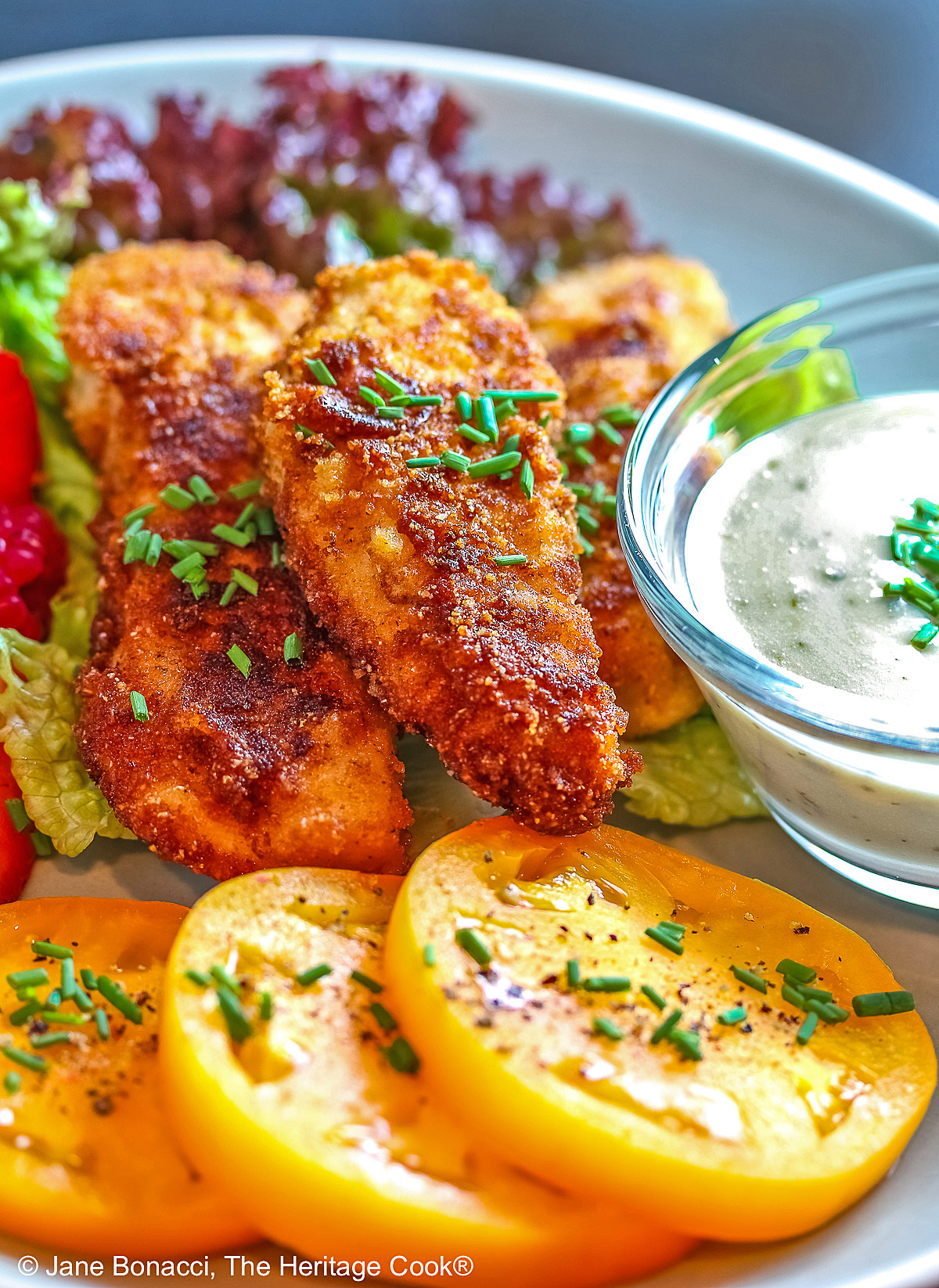 Pile of Buttermilk Fried Chicken Fingers sitting on a green lettuce leaf, on a turquoise plate surrounded by red and blue berries and slices of yellow tomato with a bowl of ranch dressing for dipping, on a black background © 2025 Jane Bonacci, The Heritage Cook. 