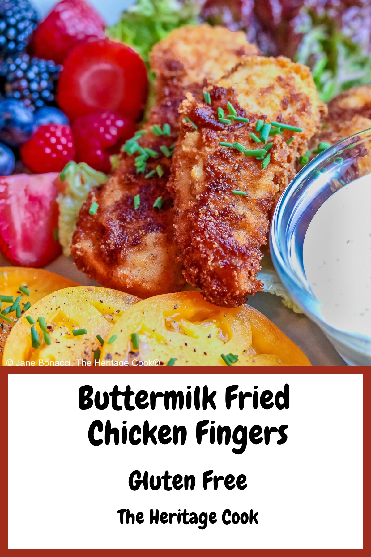 Pile of Buttermilk Fried Chicken Fingers sitting on a green lettuce leaf, on a turquoise plate surrounded by red and blue berries and slices of yellow tomato with a bowl of ranch dressing for dipping, on a black background © 2025 Jane Bonacci, The Heritage Cook.