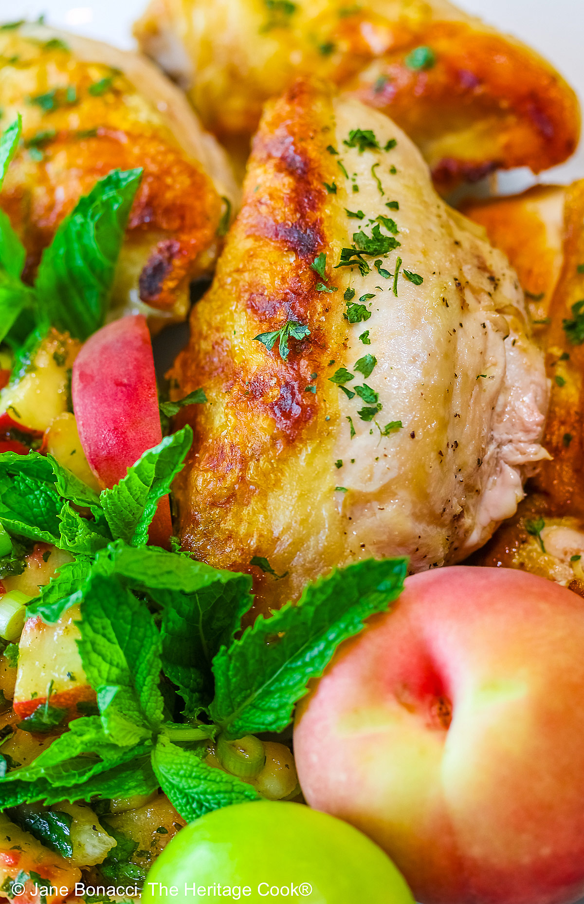 Grilled chicken breast halves piled on a white platter with the chunky peach poblano chimichurri in front with peach wedges around the edge and multiple mint sprigs tucked into the top © 2025 Jane Bonacci, The Heritage Cook. 