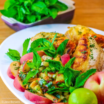 Grilled chicken breast halves piled on a white platter with the chunky peach poblano chimichurri in front with peach wedges around the edge and multiple mint sprigs tucked into the top © 2025 Jane Bonacci, The Heritage Cook.