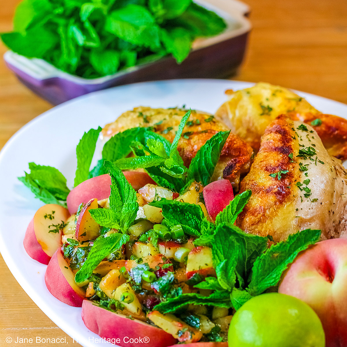 Grilled chicken breast halves piled on a white platter with the chunky peach poblano chimichurri in front with peach wedges around the edge and multiple mint sprigs tucked into the top © 2025 Jane Bonacci, The Heritage Cook.