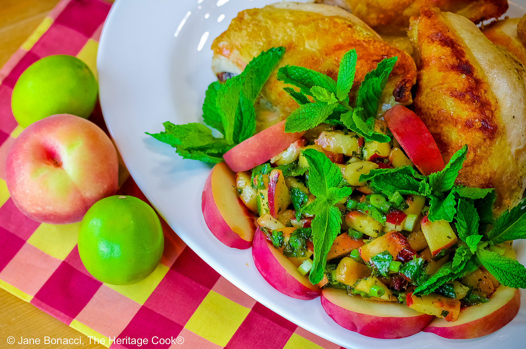 Grilled chicken breast halves piled on a white platter with the chunky peach poblano chimichurri in front with peach wedges around the edge and multiple mint sprigs tucked into the top © 2025 Jane Bonacci, The Heritage Cook. 
