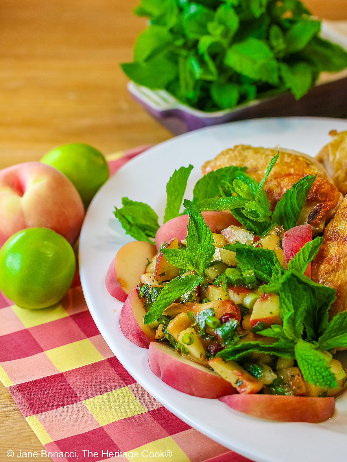 Grilled chicken breast halves piled on a white platter with the chunky peach poblano chimichurri in front with peach wedges around the edge and multiple mint sprigs tucked into the top © 2025 Jane Bonacci, The Heritage Cook. 