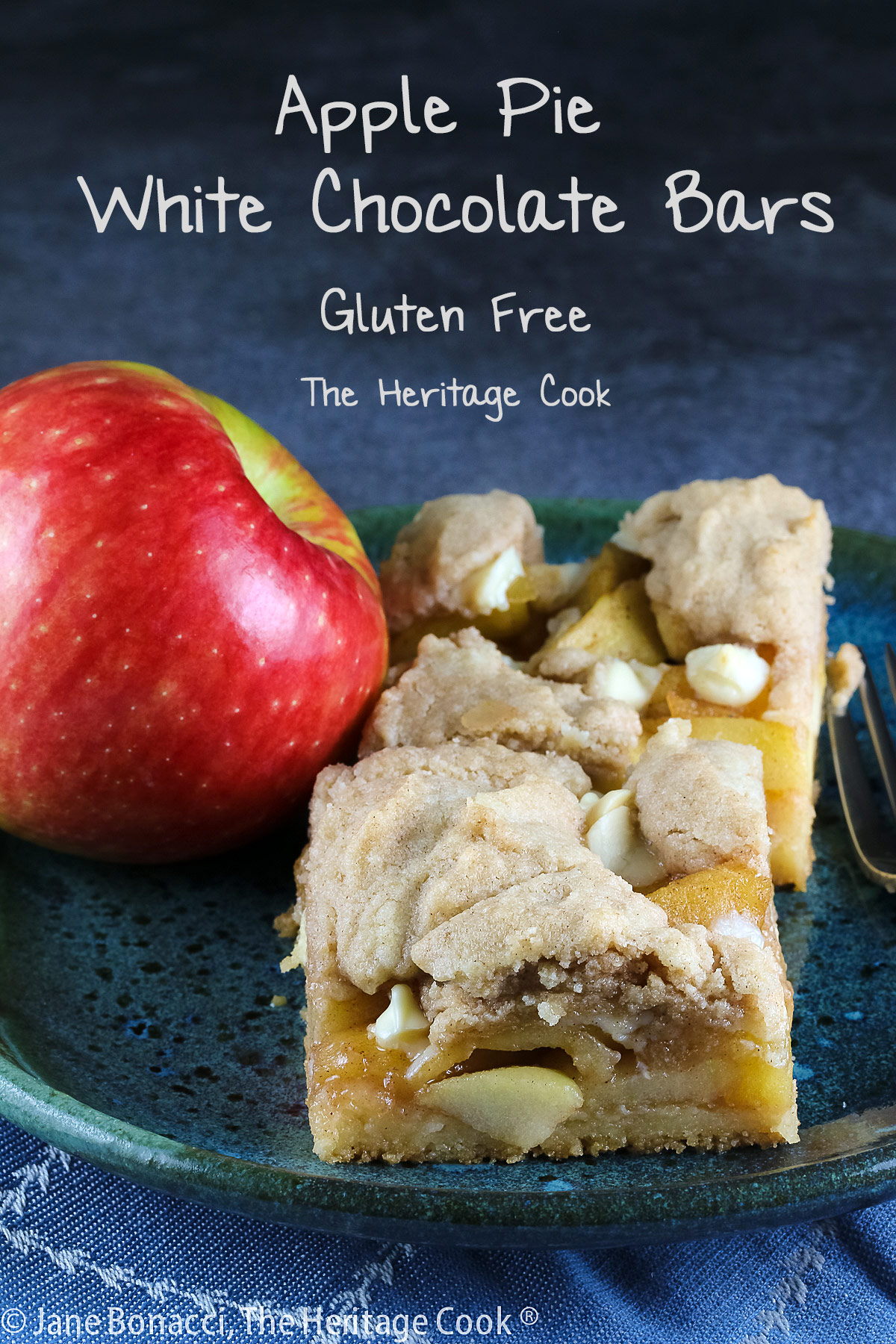 2 squares of Apple Pie White Chocolate Bars on a teal colored plate sitting on a blue cloth with a red apple on the plate or just behind © 2025 Jane Bonacci, The Heritage Cook. 