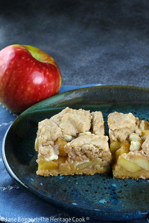 2 squares of Apple Pie White Chocolate Bars on a teal colored plate sitting on a blue cloth with a red apple on the plate or just behind © 2025 Jane Bonacci, The Heritage Cook. 