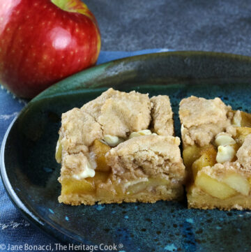 2 squares of Apple Pie White Chocolate Bars on a teal colored plate sitting on a blue cloth with a red apple on the plate or just behind © 2025 Jane Bonacci, The Heritage Cook.