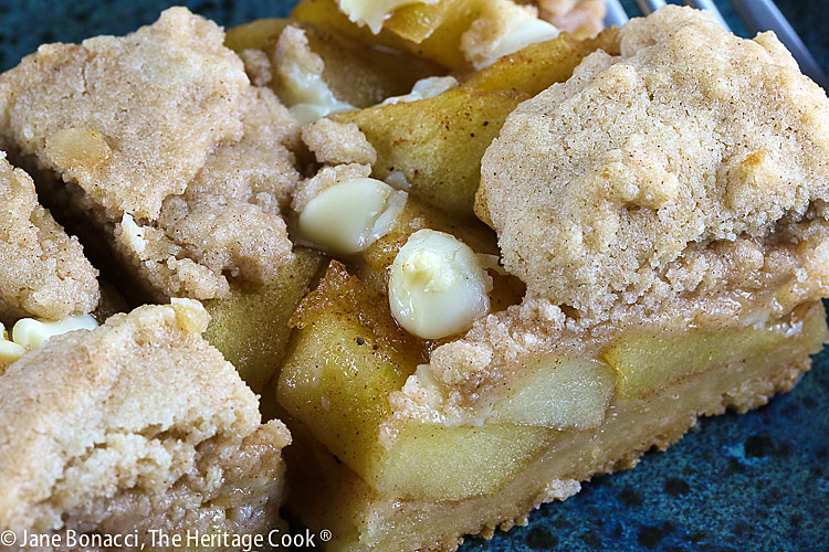 2 squares of Apple Pie White Chocolate Bars on a teal colored plate sitting on a blue cloth with a red apple on the plate or just behind © 2025 Jane Bonacci, The Heritage Cook. 