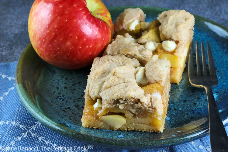 2 squares of Apple Pie White Chocolate Bars on a teal colored plate sitting on a blue cloth with a red apple on the plate or just behind © 2025 Jane Bonacci, The Heritage Cook. 