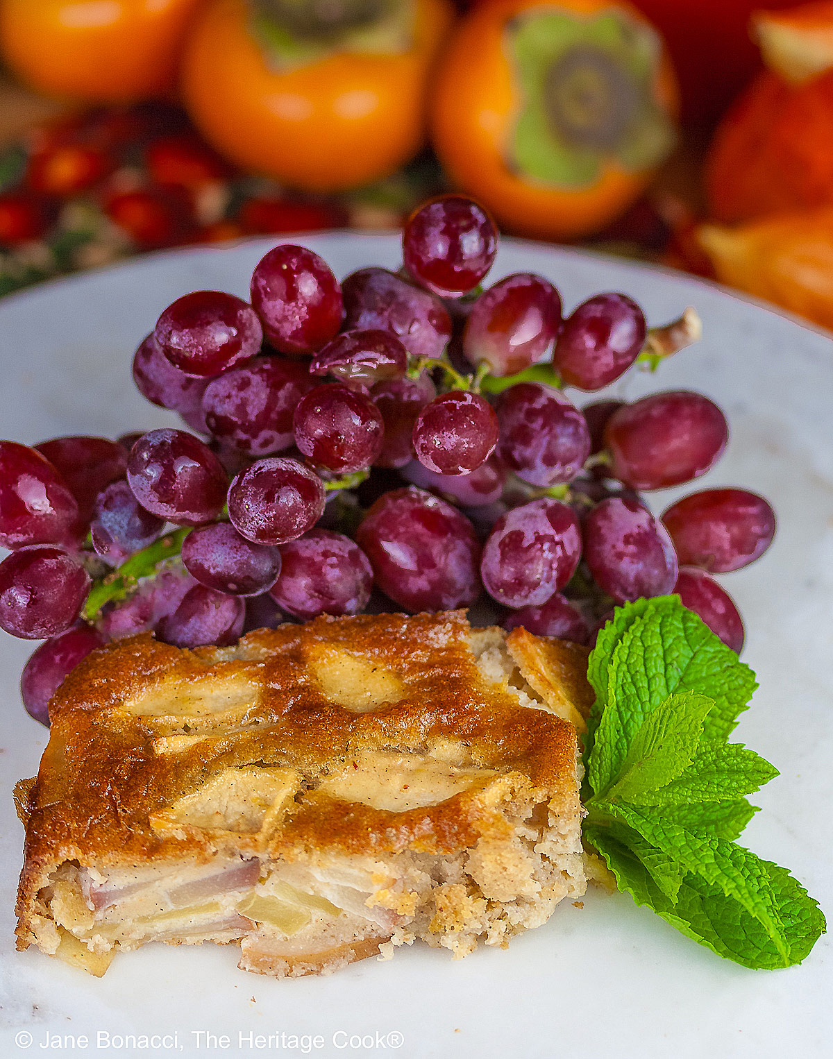 A gorgeous browned square cake with tons of fruit breaking the surface on a round white marble surface with red and orange fall fruits behind, a slice with a sprig of mint and red grapes flanking it © 2026 Jane Bonacci, The Heritage Cook.